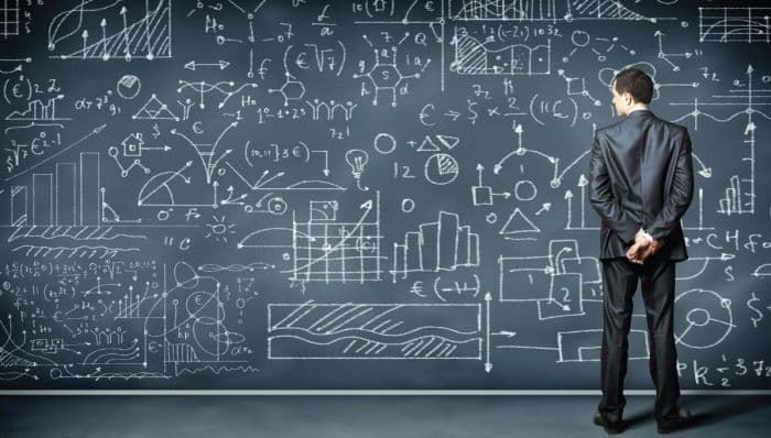 a businessman stands in front of a giant blackboard filled with chalk drawings of graphs, charts, and formulas
